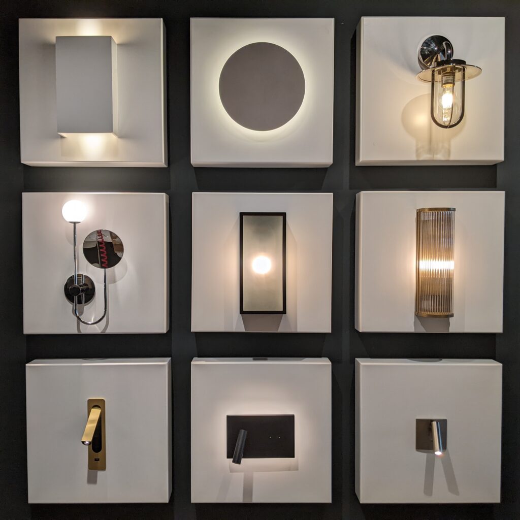 A grid of nine different modern wall lights displayed on a dark background. Each light has a unique geometric design and emits a soft glow, emphasizing shapes like circles, rectangles, and cylinders.