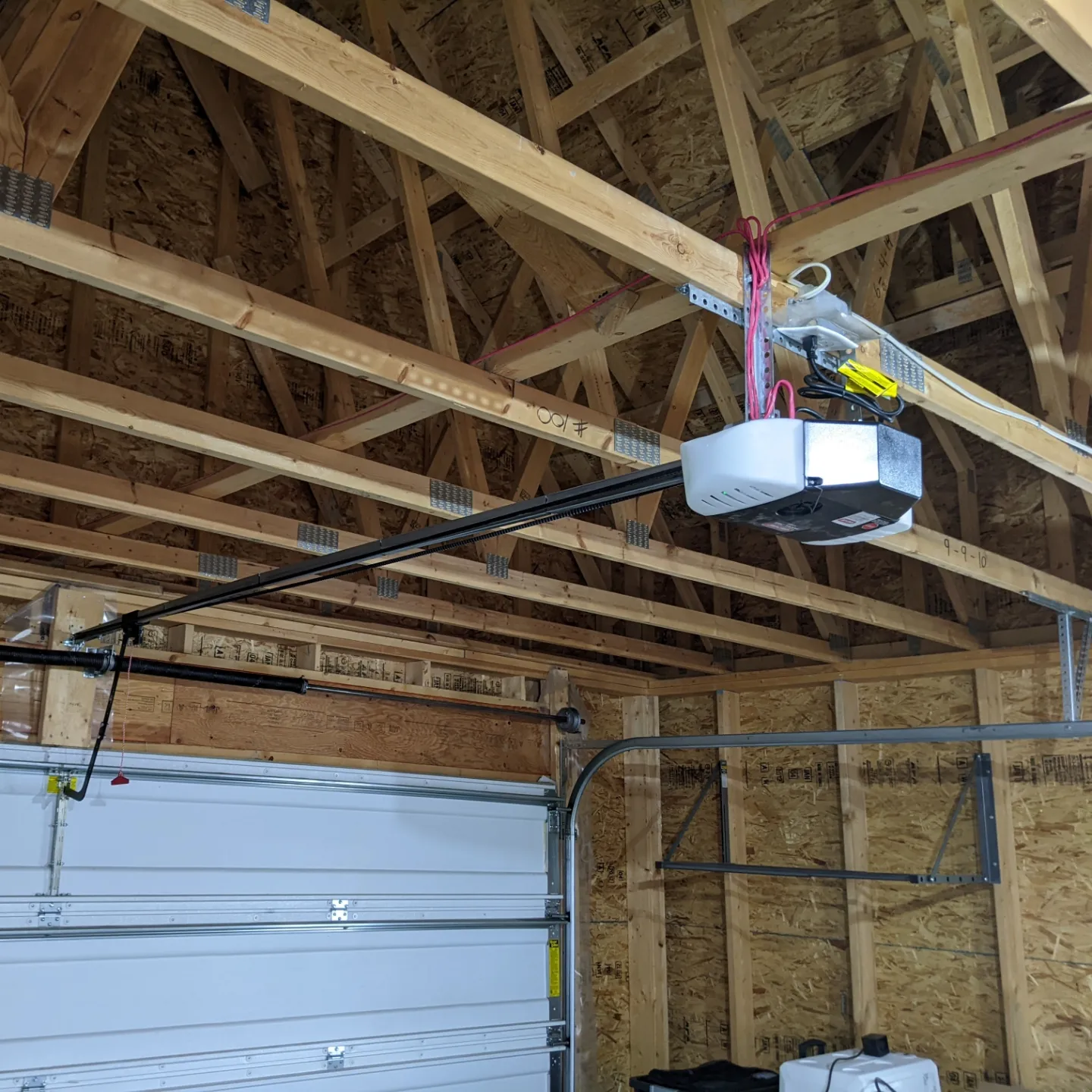 A garage interior with an unfinished wooden ceiling. A white automatic garage door opener is mounted, with a red pull cord attached. The garage door is partially visible, and the walls are made of bare wood panels.