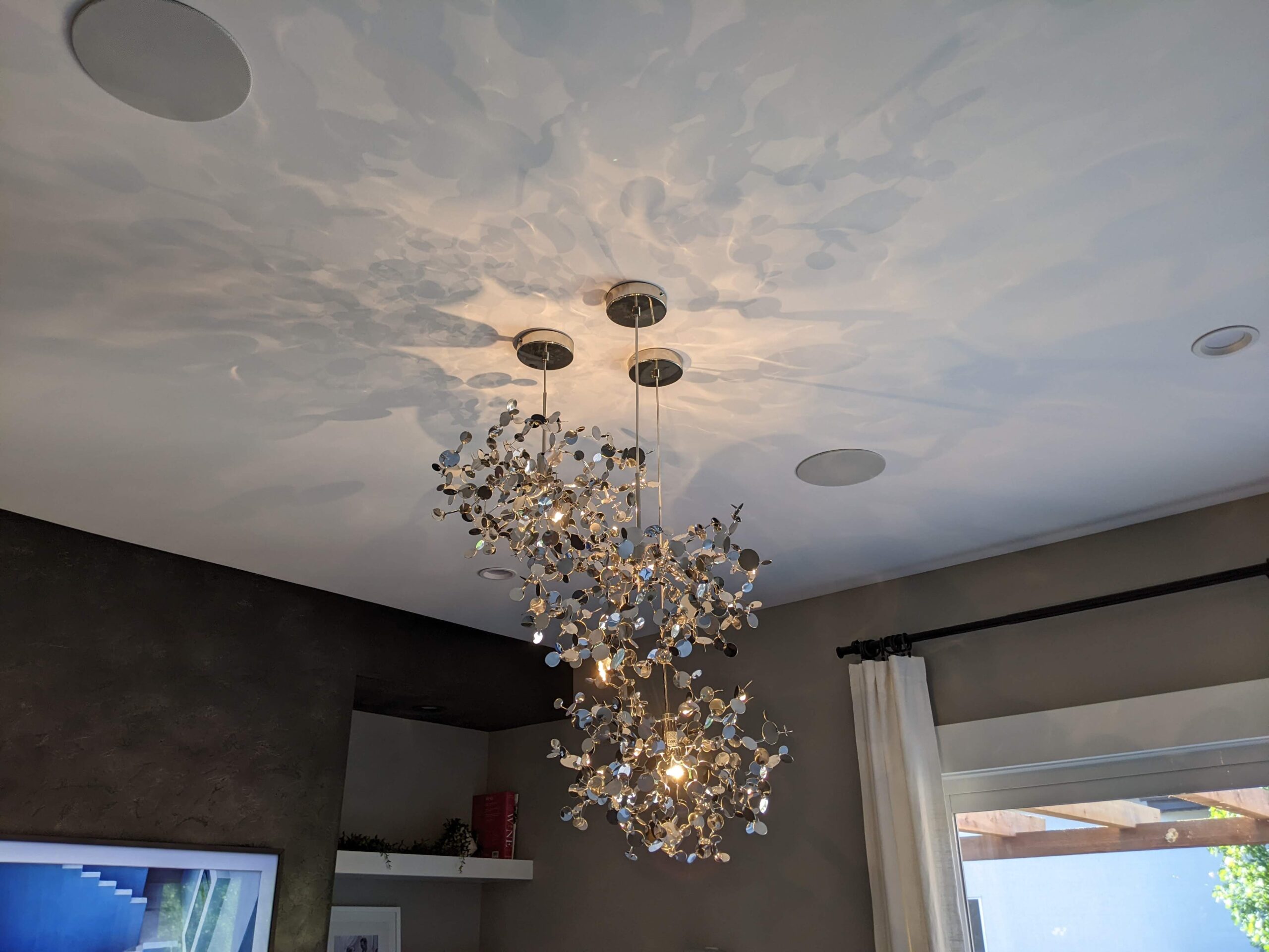 A modern ceiling light fixture with glass and metal elements creates a reflective pattern on the ceiling. It hangs in a room with light gray walls and a partially visible window with a white curtain.