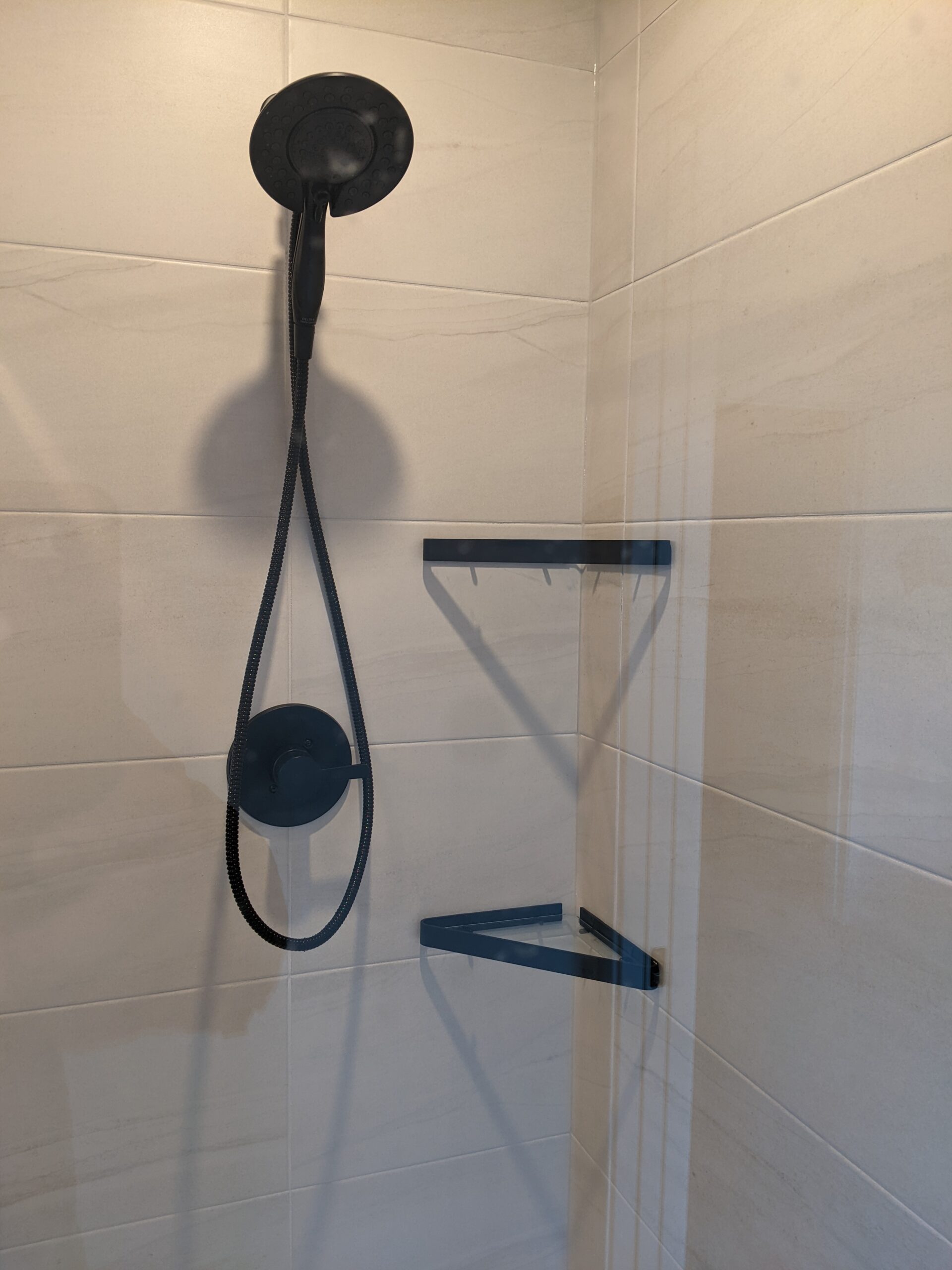 A shower with a black showerhead and hose is mounted on a tiled wall. There are two triangular black shelves attached to the corner, casting shadows on the light beige tiles.